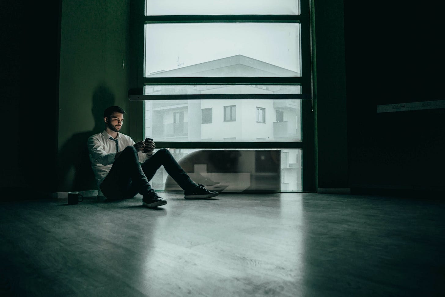 A photo of a sad-looking man sitting on the floor in a dark room checking his phone. A photo of a sad-looking man sitting on the floor in a dark room checking his phone.