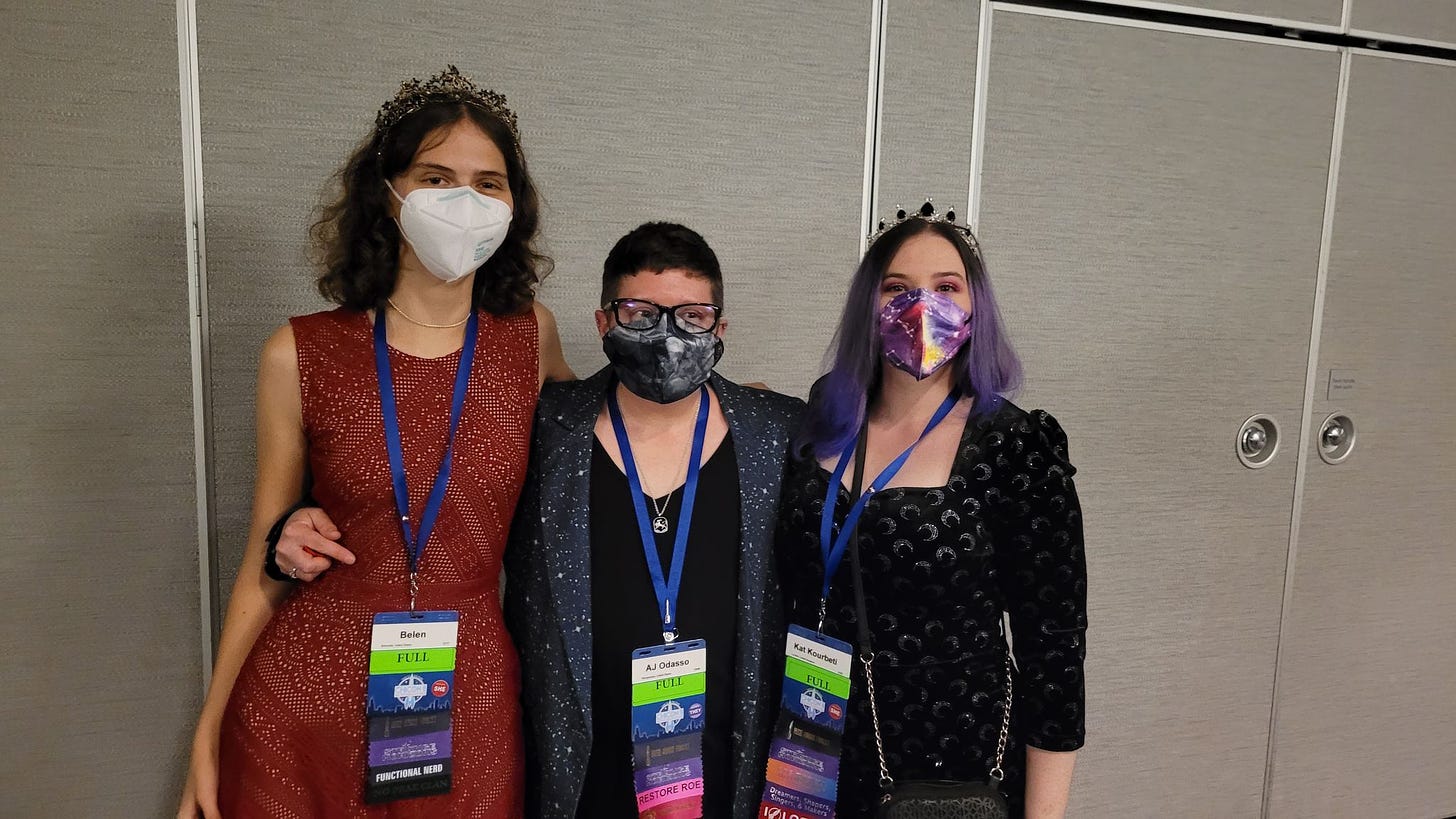 a photo of the three strange horizons representatives at the hugo awards reception in chicago last september. left to right: belen edwards, first reader, a tall brunette in a red dress and gold tree branch tiara, wearing a white n95 mask and her worldcon name badge; a.j. odasso, poetry editor, with short brown hair, in a star-print blazer, glasses, and a black n95 mask, also sporting a silver necklace and their worldcon name badge; myself, kat kourbeti, in a moon-print velvet bodycon dress, wearing a silver and black tiara on top of my long purple hair, a galaxy-print n95 mask, and my worldcon name badge.