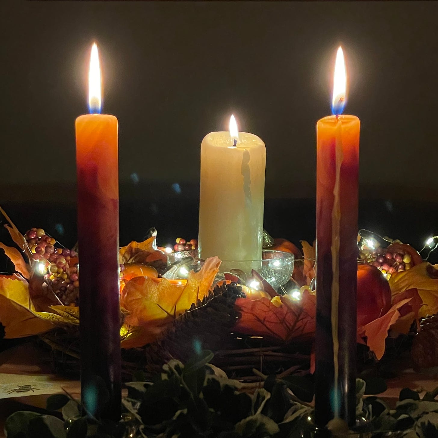 Photo of three lit candles surrounded by leaves and greenery and berries and shining glass. The candles are clearly the only light in the room.