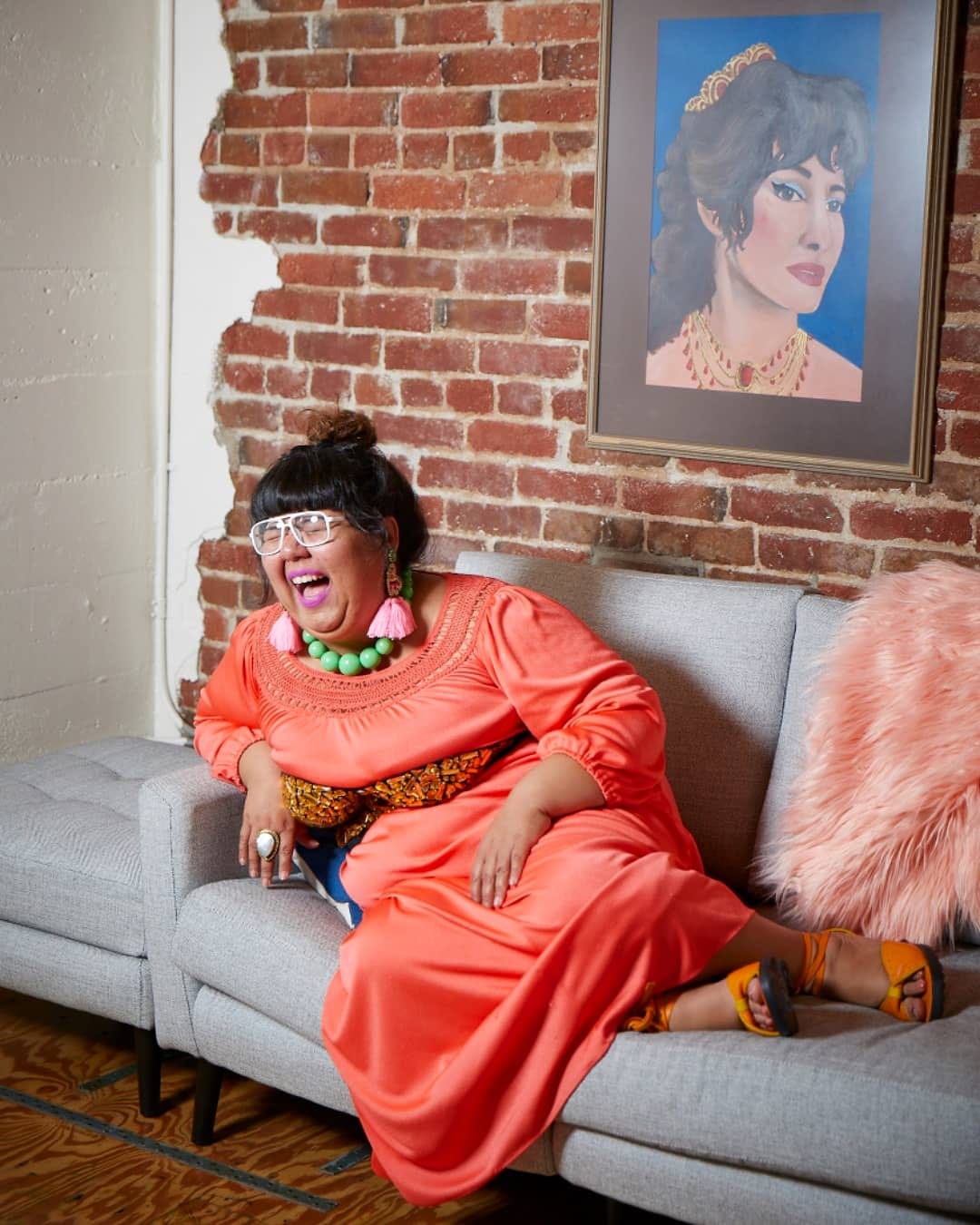 Today's guest Virgie Tovar, laughing while sat on a sofa against an exposed red brick wall