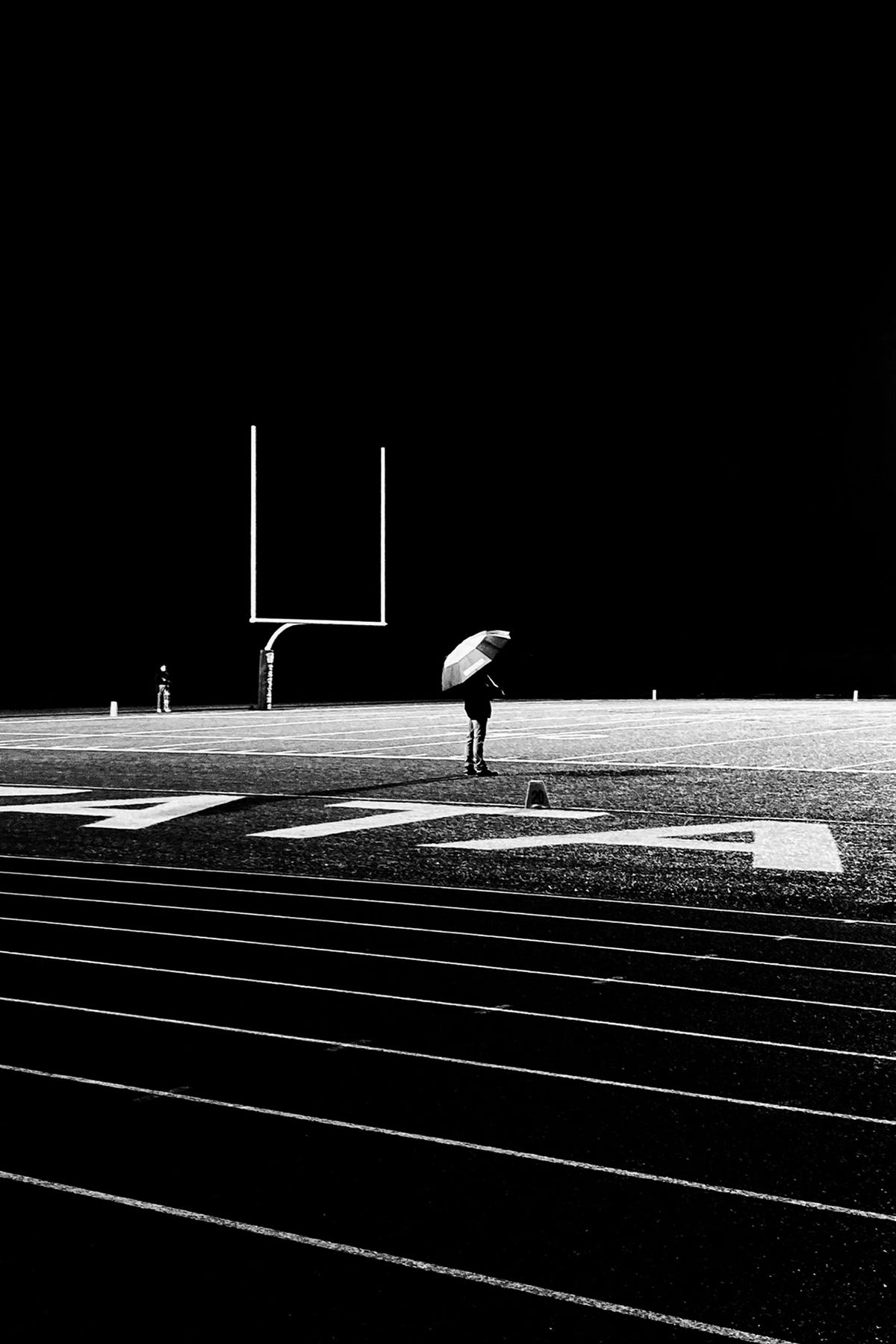 A man with an umbrella stands on an empty part of a football field in the rain. 