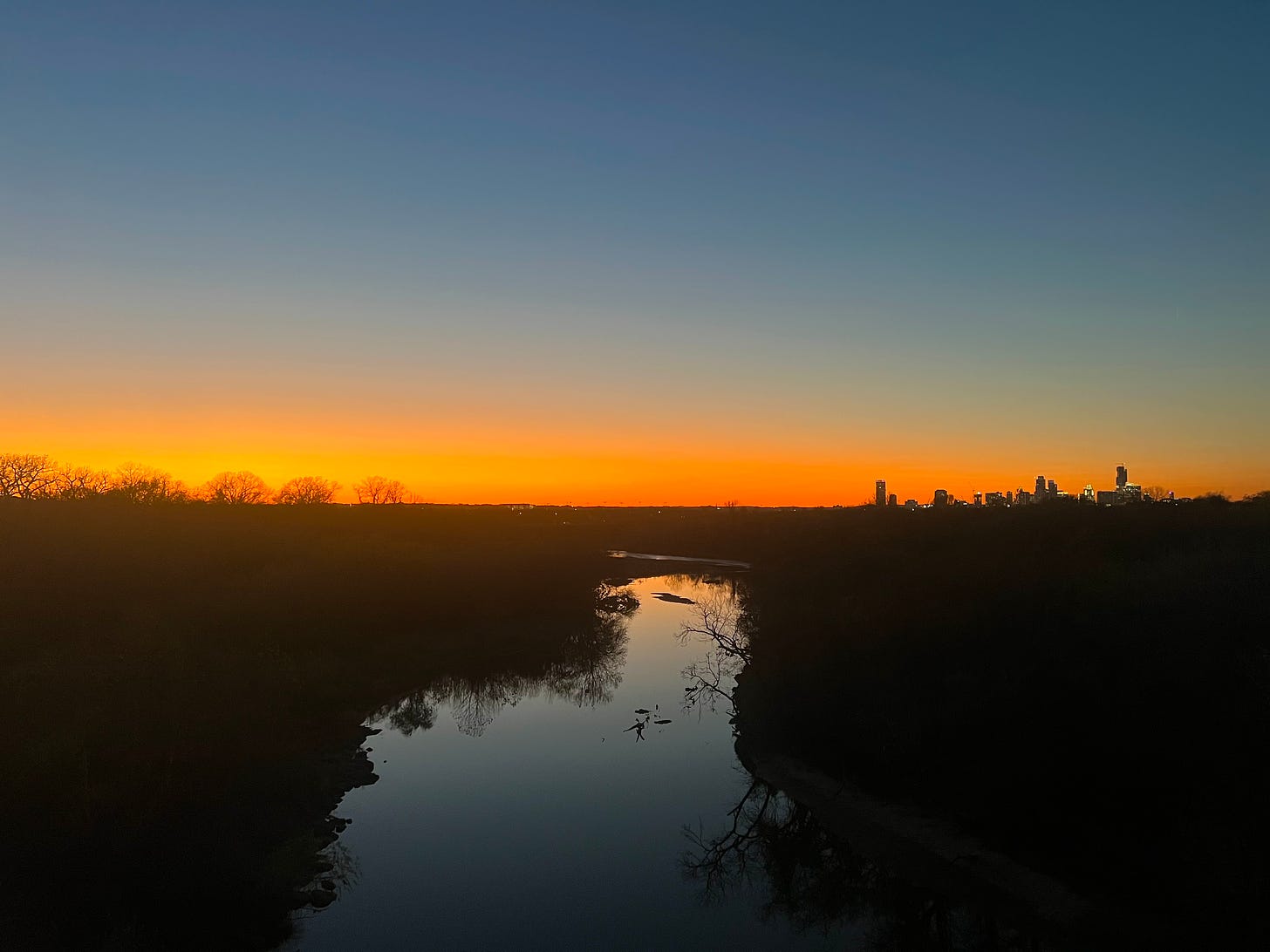 Sunset over Austin, with wild urban river in the foreground and the skyline of downtown silhouetted on the horizon