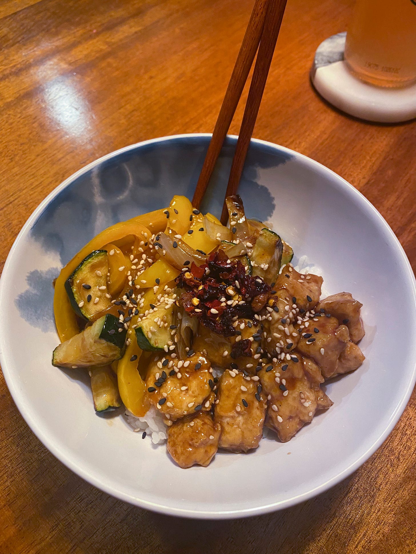 In a white bowl with wooden chopsticks sticking out at the back, white rice is topped with pieces of tofu and stir fried zucchini and pepper in teriyaki sauce. On top are black and white sesame seeds and chili crisp.