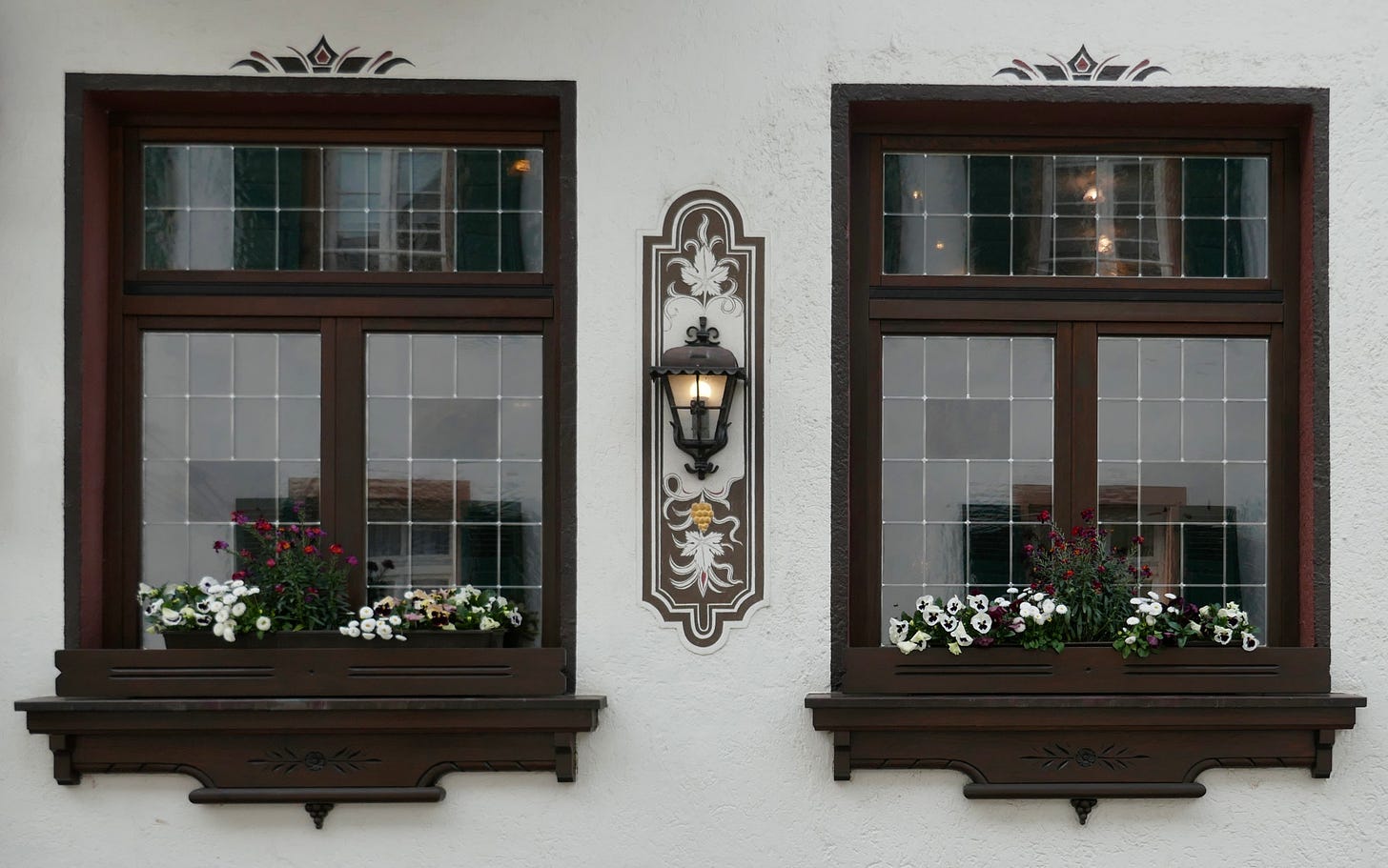 two identical windows against white stucco wall with flower boxes of pansies and other flowers with a lighted sconce between them