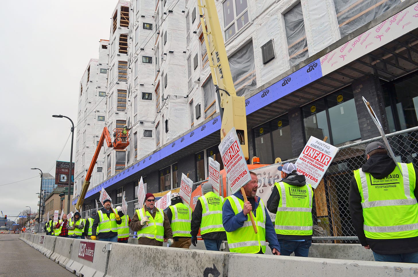 workers wearing neon yellow vests carry picket signs reading "unfair" in front of a building under construction