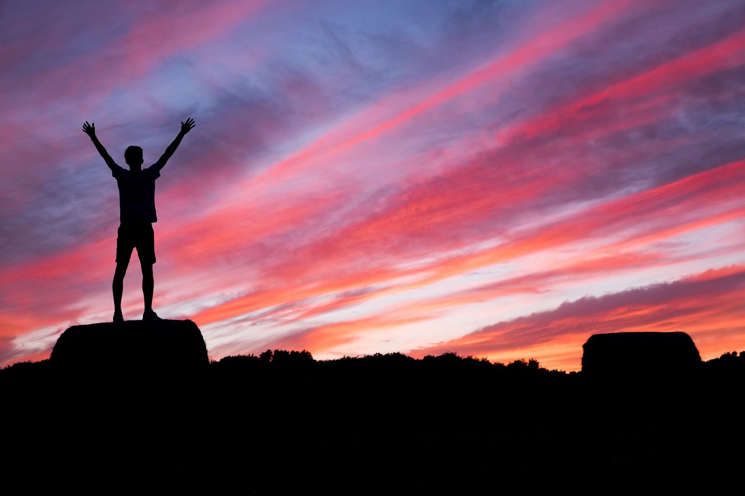 A man raises his arms in triumph while standing on a large rock and looking at a colorful sunset
