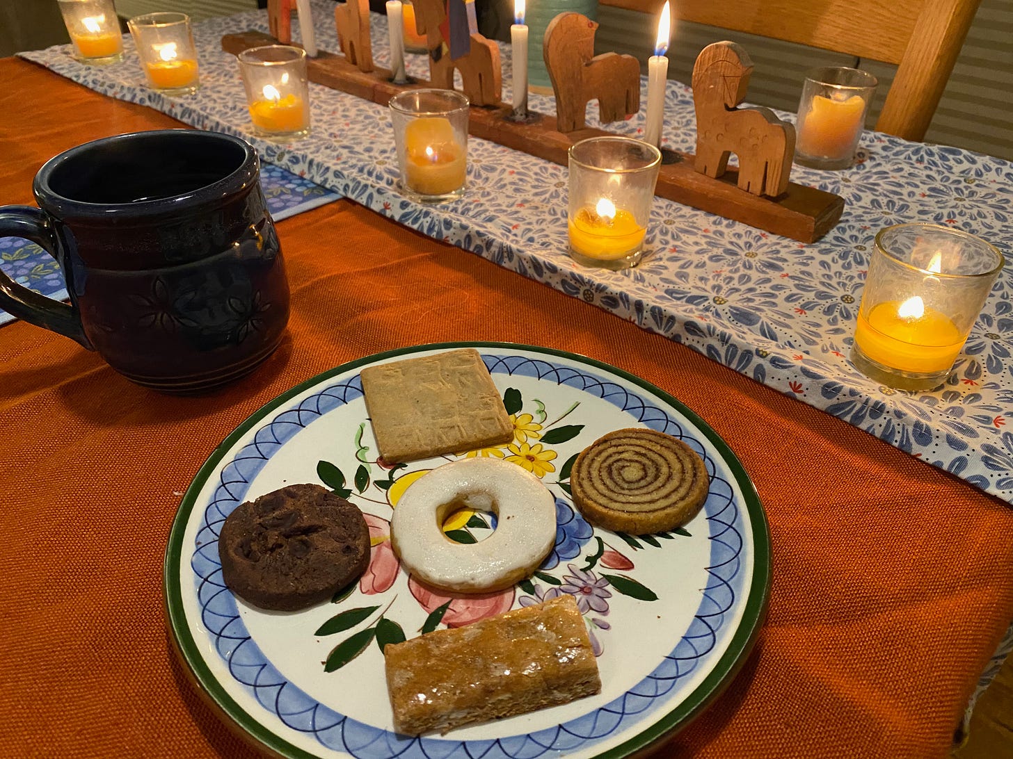 A teatime scene. The table is covered with a red tablecloth. A Swedish advent candle holder is lit, as are several smaller beeswax candles in small glass cups. There is a blue ceramic mug next to a plate that holds five cookies of different shapes and colors.