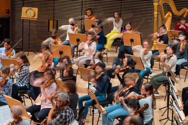 Violinists and harpists, most of whom are women, play at a recent rehearsal on the stage of David Geffen Hall. All the players are wearing street clothes.