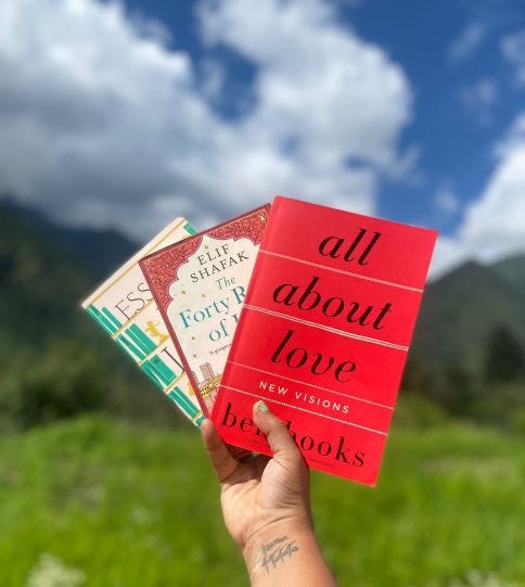 Image of me holding three books up in front of mountains and blue sky with white clouds. the books are from left to right: Essays about Love (Alain de Botton), The Forty Rules of Love (Elif Shafak) and all about love new visions (bell hooks)