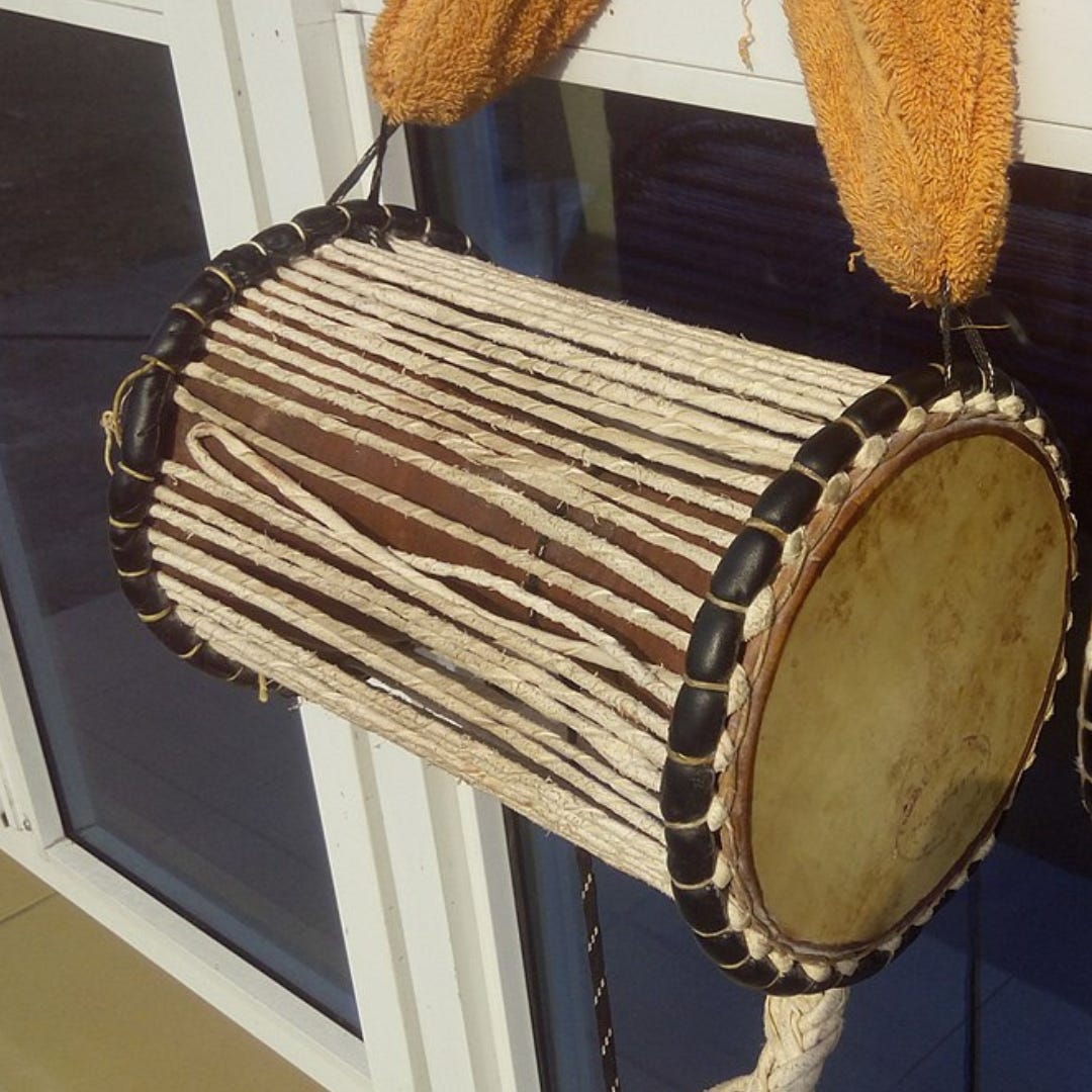 A talking drum from Western Nigeria is hung from its sling. The medium-size drum has vertical rope woven around it and thick leather wrapped horizontally around the top and bottom of it. 