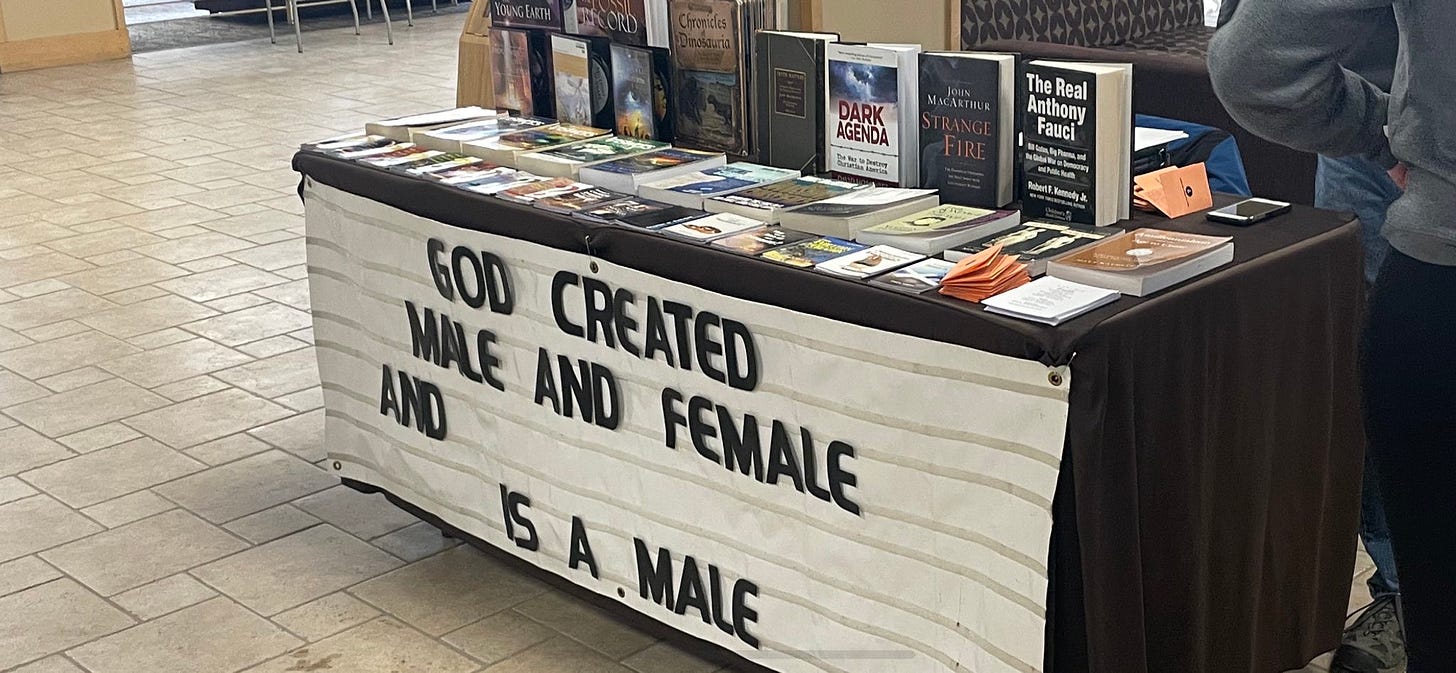 Schmidt's table is covered with biblical and conspiratorial books. The banner hanging from the front reads "God created male and female and [blank] is a male."