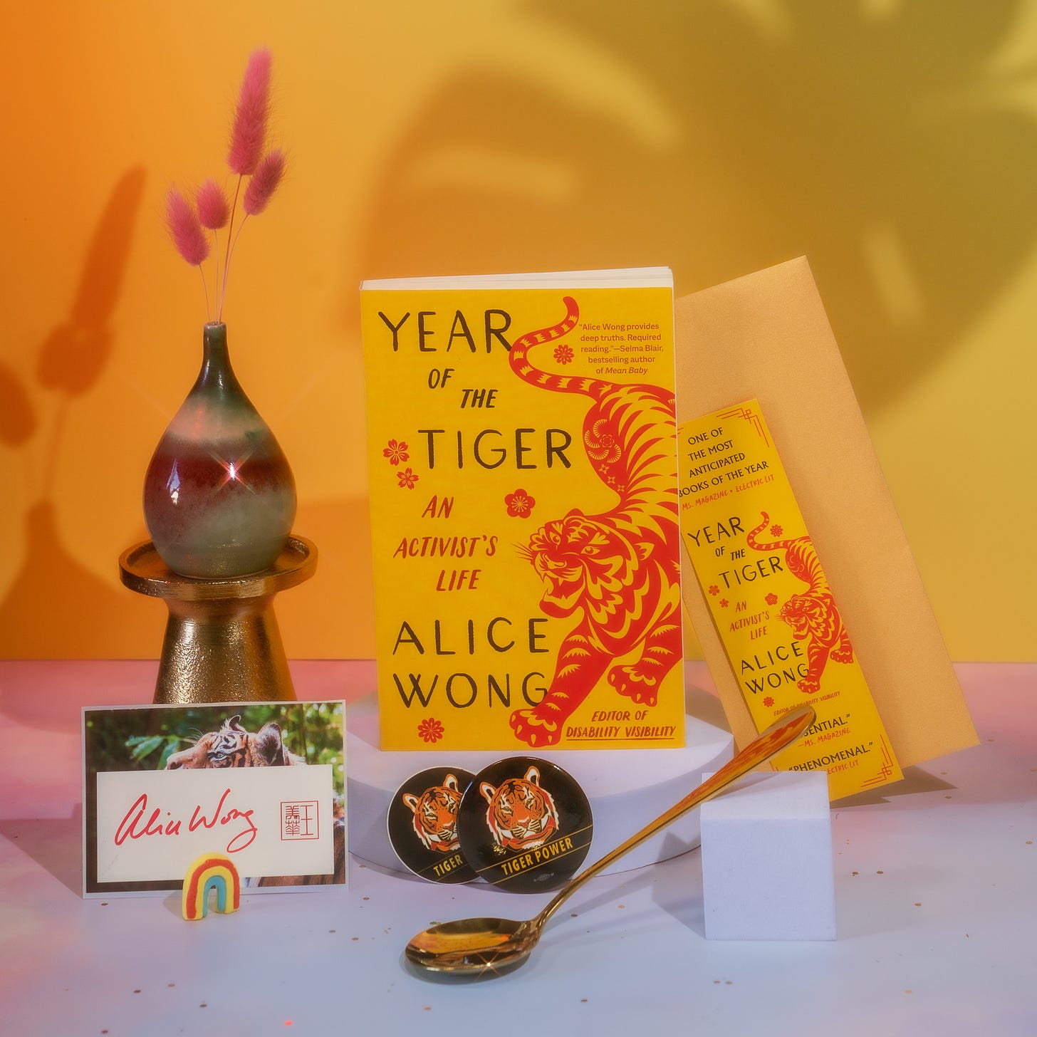 Photo with a tangerine background with a pink border at the bottom with a copy of Year of the Tiger paperback in the center. A tiger bookmark positioned to the right of the book. On the left is a multicolored pear-shaped vase holding pink floral decorations. A signed tiger bookplate by Alice Wong in front of the vase with a small rainbow decor standing upright. In the center is one round tiger sticker and one round tiger button with a gold dipper spoon from @umeshiso_ positioned horizontally at an angle. A tiger bookmark positioned to the right of the book. Photo credit: @ziru.mo (on IG)