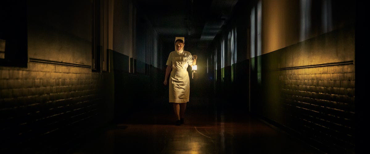 A nurse in a 1970s uniform walking down a dark hospital hallway with a lamp. She is centered in the frame, and the only source of light is from her lamp and some from the windows behind her.