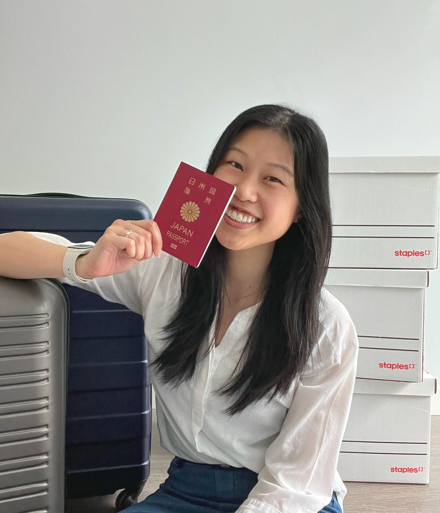 a girl with long black hair, wearing a white shirt and jeans. she is holding a red japanese passport in her hand in front of two suitcases and boxes