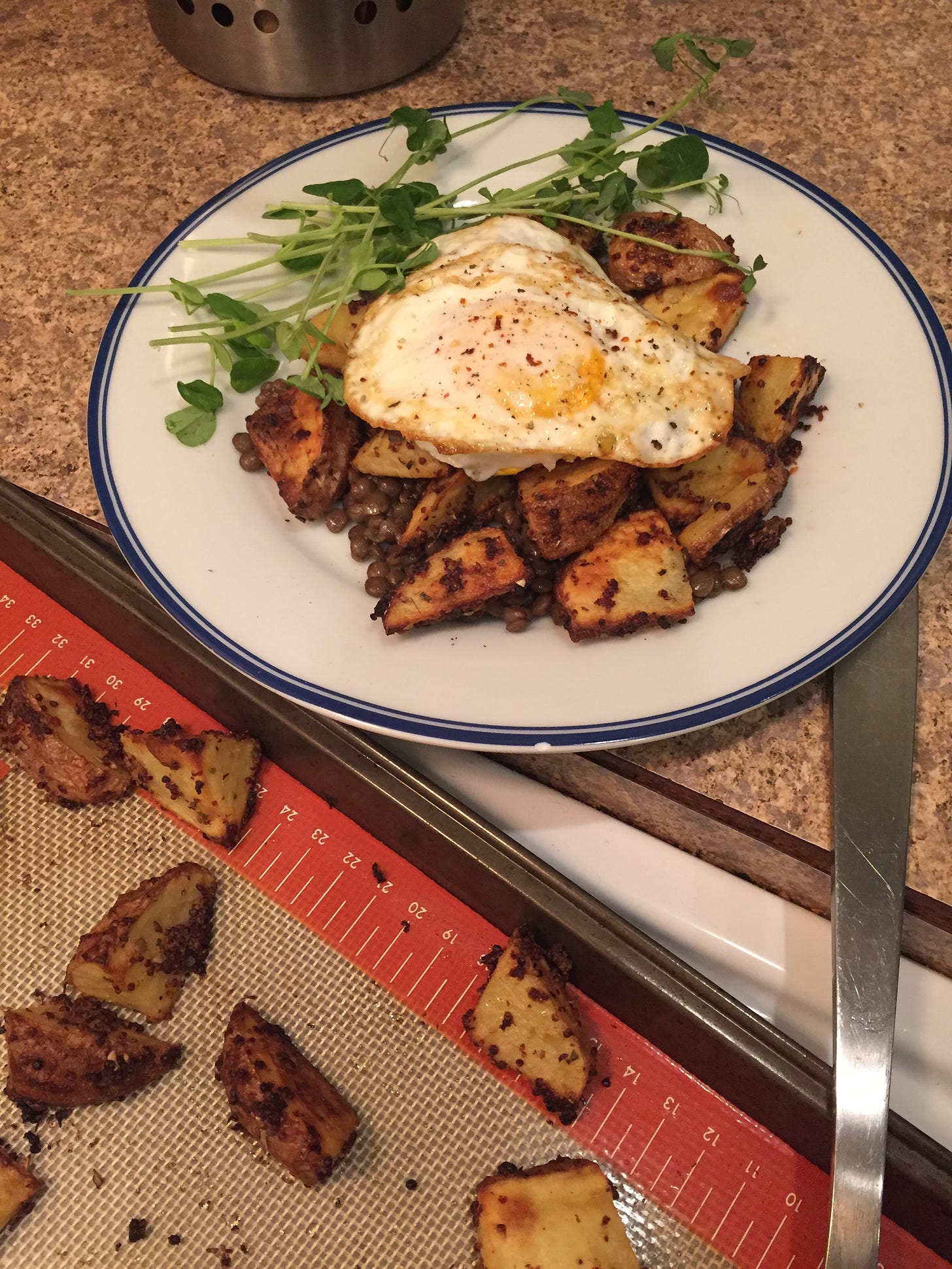 In the foreground, a corner of a baking pan covered with a silpat and pieces of potato, and the handle of a metal spatula is just visible. Behind it is a plate holding some of the potatoes on top of a bed of lentils, with a crispy fried egg sprinkled with chili flakes on top, and a little pile of pea shoots to the side.
