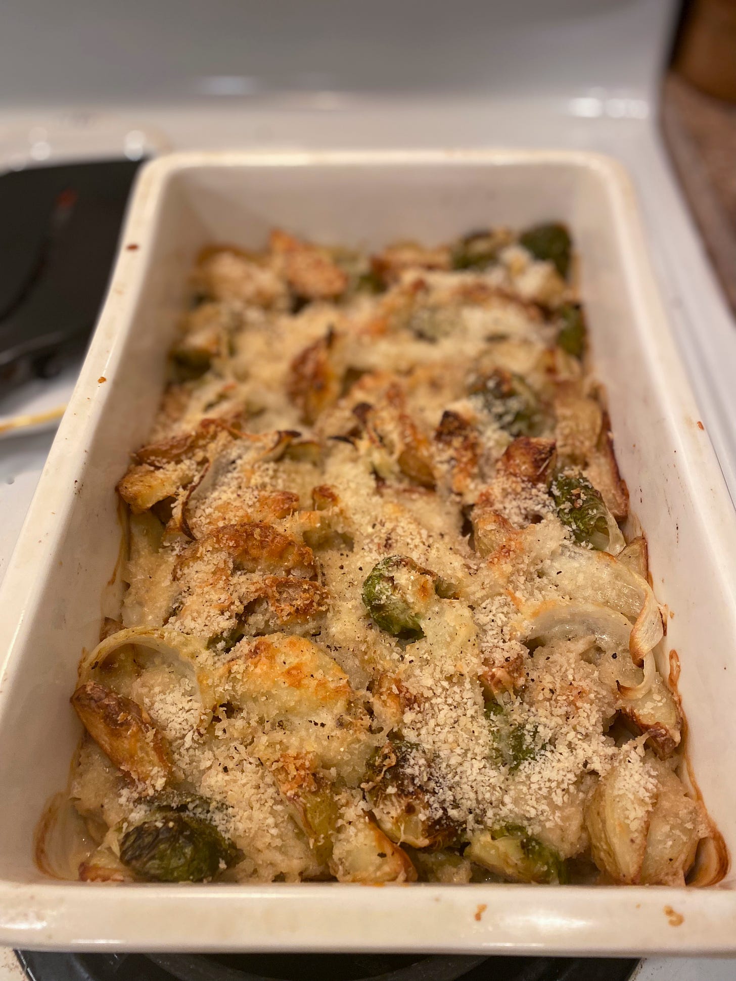 A white rectangular baking dish on top of the stuff, full of roasted wedges of potatoes and brussels sprout halves covered in cheese and breadcrumbs.