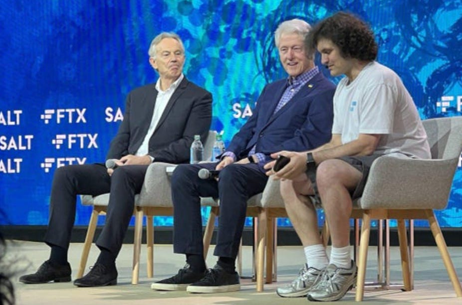 A color picture. 3 white men sit on chairs on a stage. The 2 older white men are dressed in suits and holding microphones. The younger white man is dressed in a tshirt and shorts and is wearing a microphone headset. Behind them is a blue background with "FTX" and other letters emblazoned on it in a horizontal pattern.