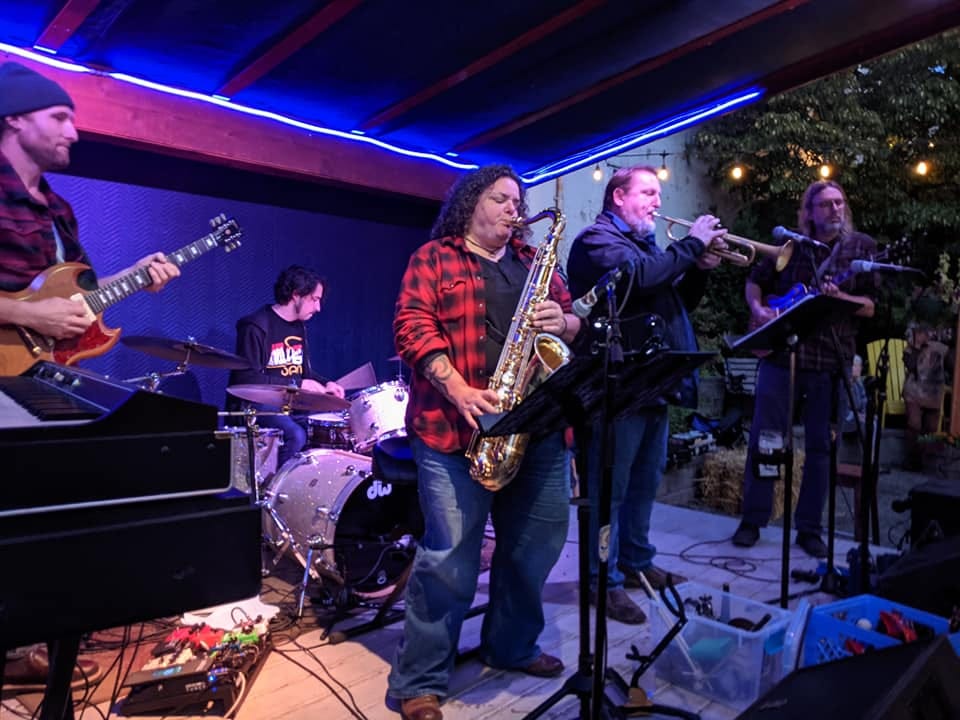 A band consisting of sax, trumpet, guitar, bass, and drums, onstage at the beer garden at Boundary Bay 