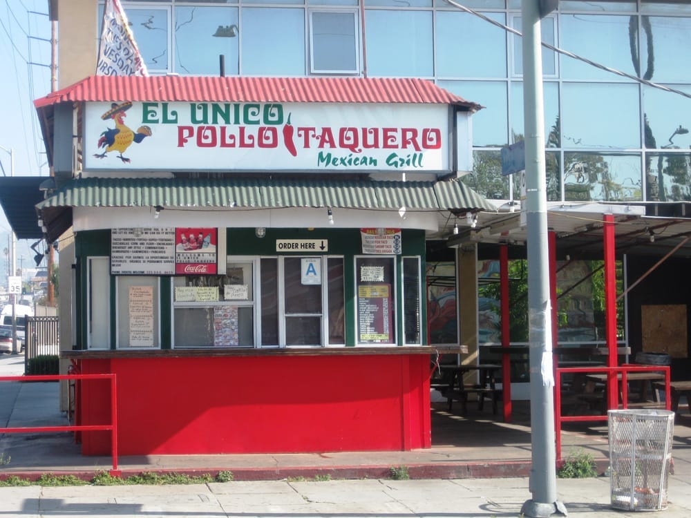 A front facing image of El Unico Pollo Taquero. The bottom part of the restaurant is red, then there are windows in between panels of green, then the sign for the business is on top. Behind the shack is a big building with glass windows. The restaurant is not open for business in this photo. 