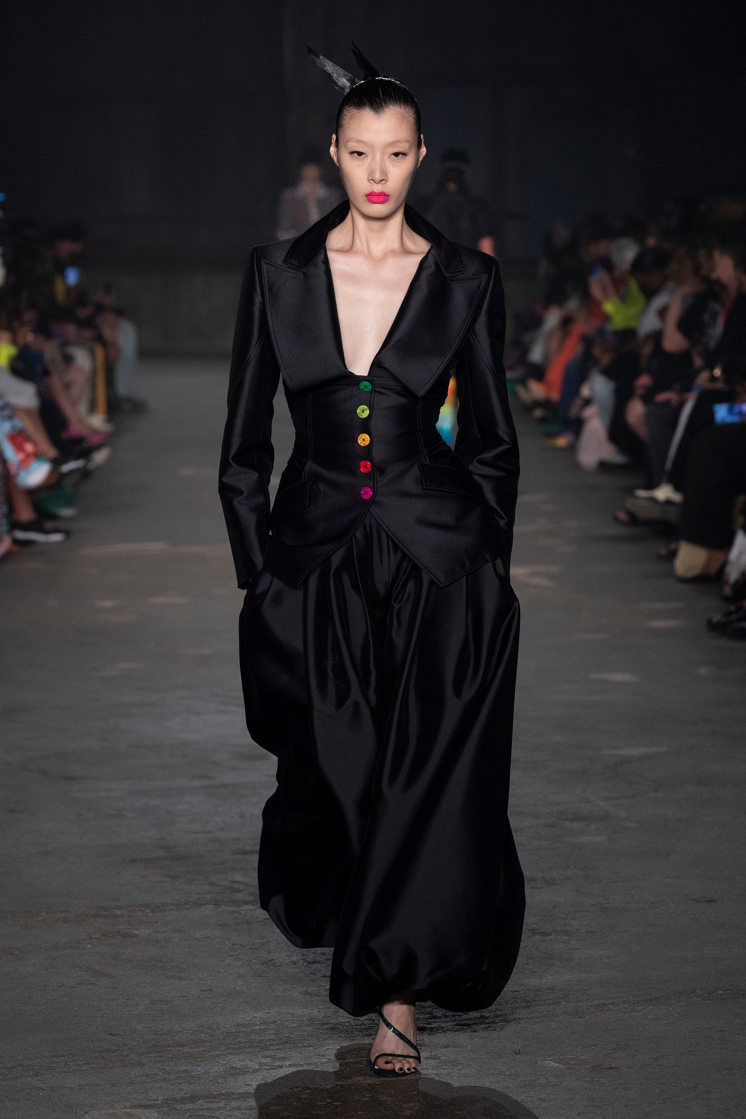 an image from Christopher John Rogers' Resort 2023 collection featuring a model wearing a black suit with rainbow buttons on the cinched waist and billowy trousers.