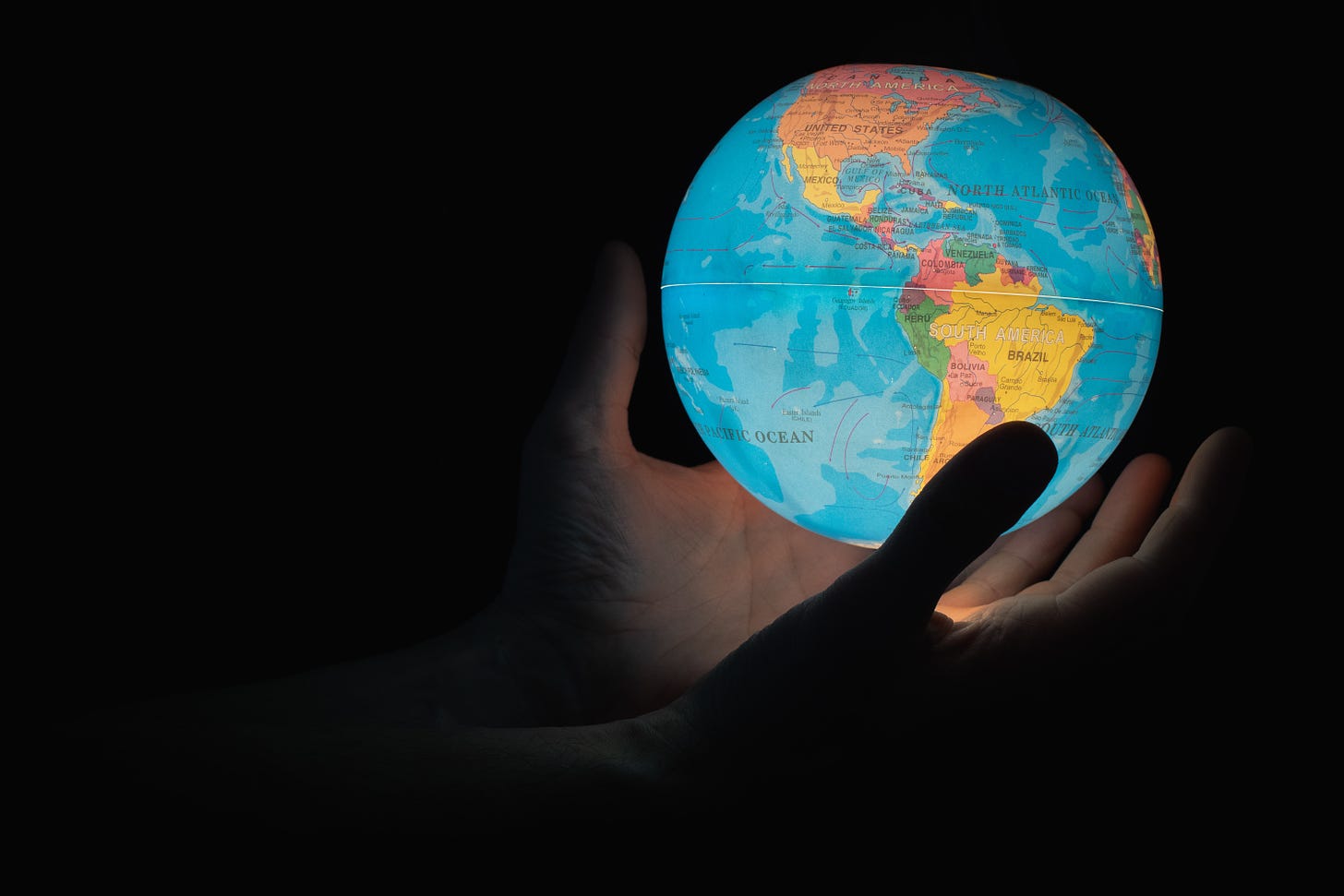 A glowing globe, illuminating two hands holding the globe against a black background. The globe is the kind you'd find in a class room, showing North and South America, each country a different color