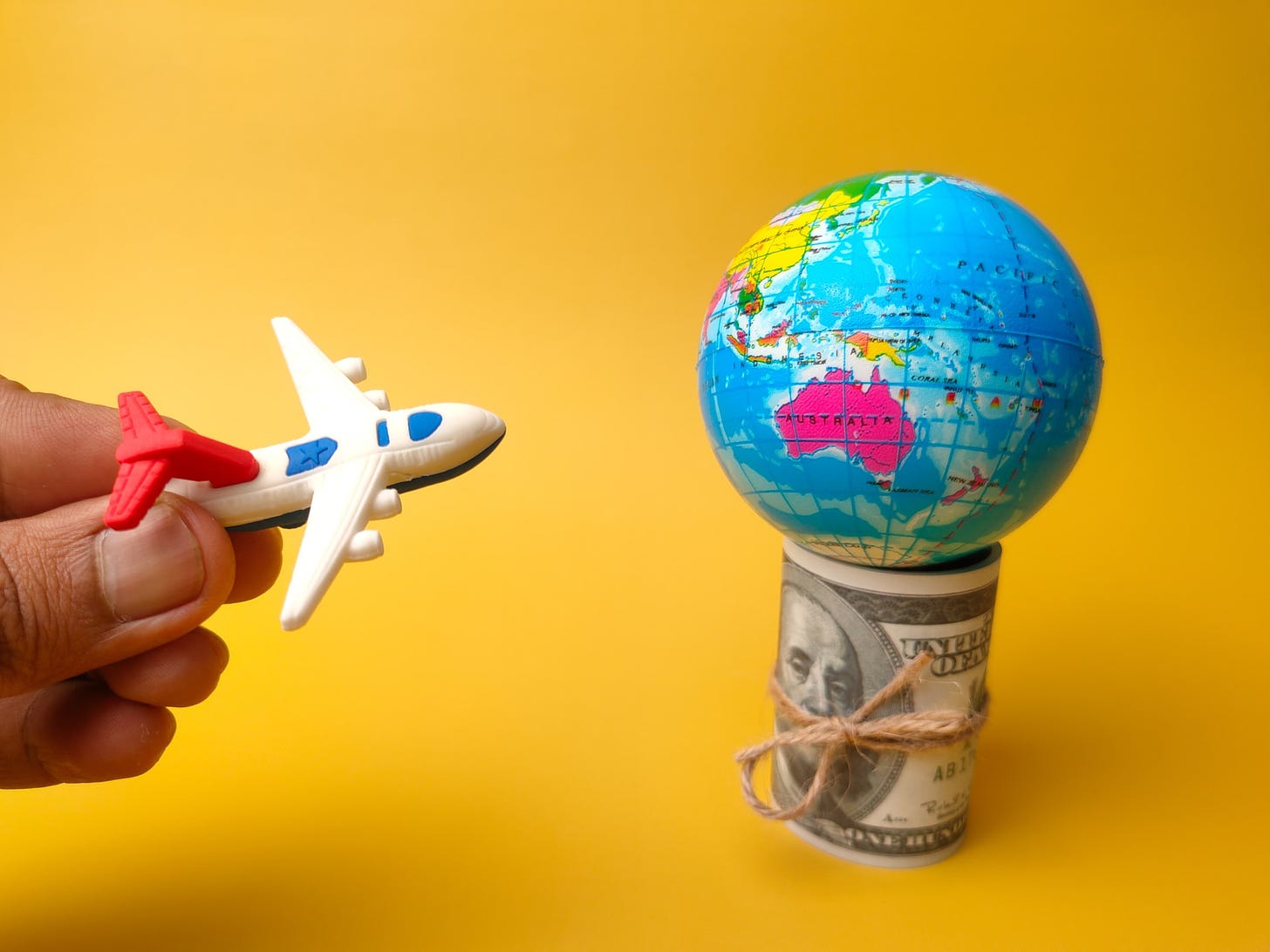 Foto de avião de brinquedo na mão de uma pessoa se aproximando de um pequeno globo colocado em cima de um maço de dinheiro, em um fundo amarelo.