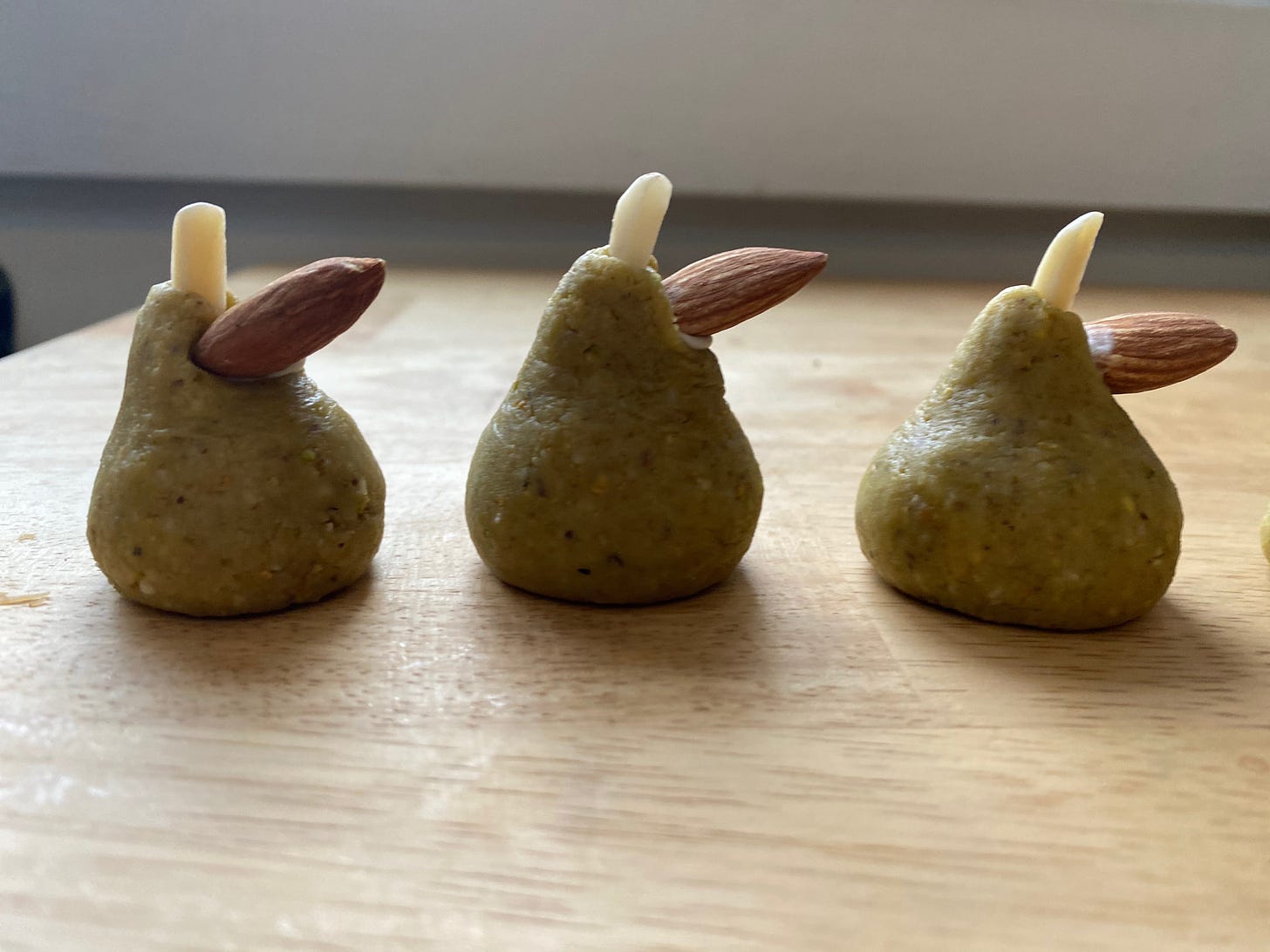 Three small pistachio marzipan pears sit on a wooden counter. They have slivered almonds sticking out of their tops to represent the pear’s stem, and a whole almond pointing out an an angle right below, to represent a leaf.