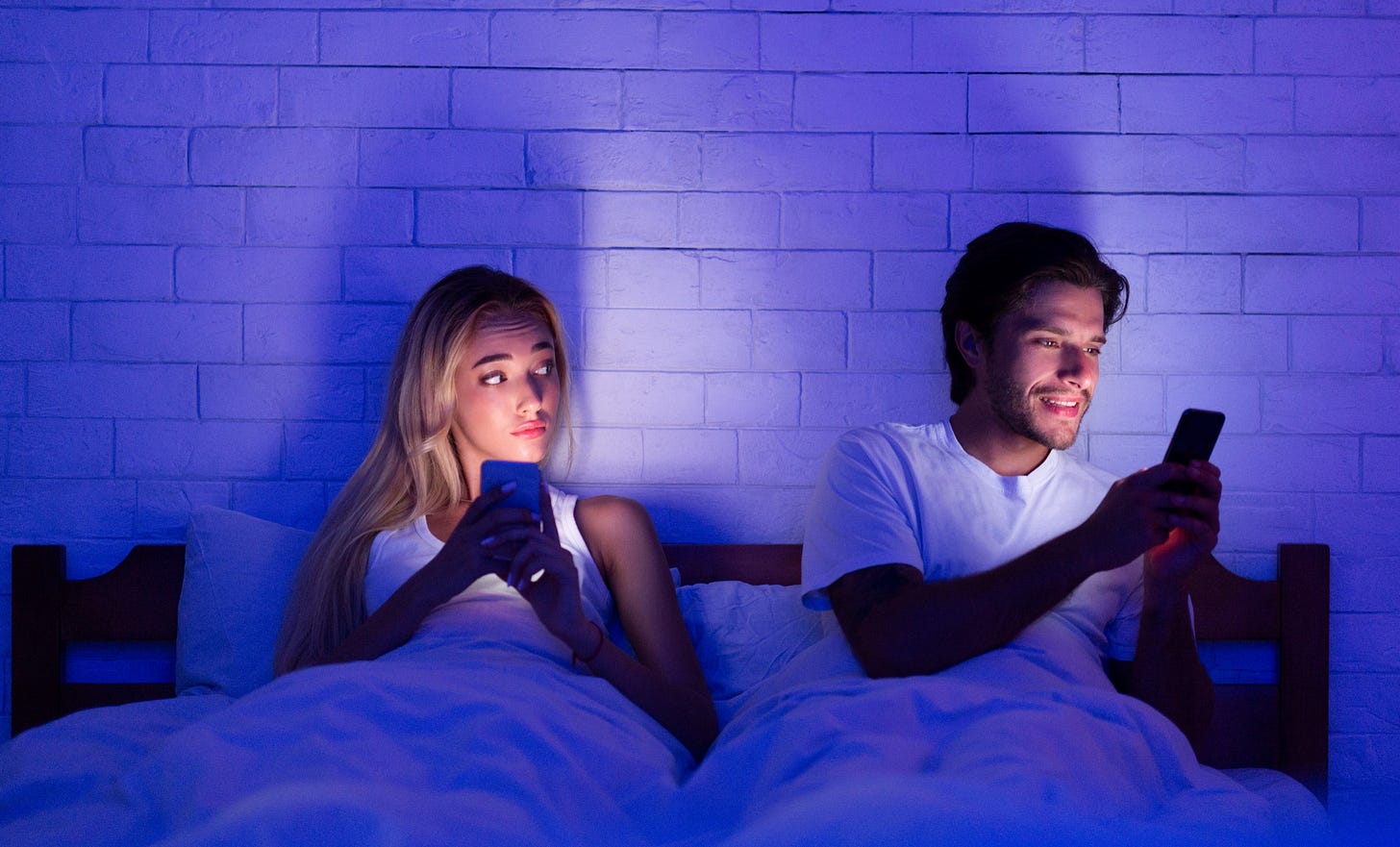 A couple lays in bed with their phones. One partner texts sneakily to the side while the other watches suspiciously.