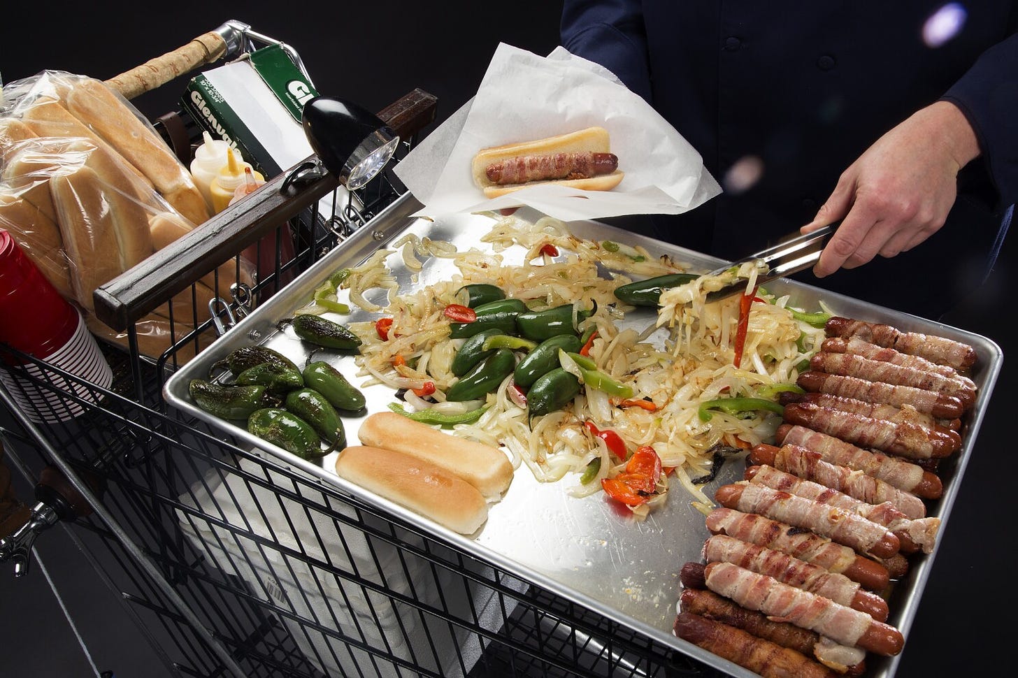 Image: A vendor grills bacon-wrapped hot dogs, peppers, and onions on a makeshift grill made out of a shopping cart