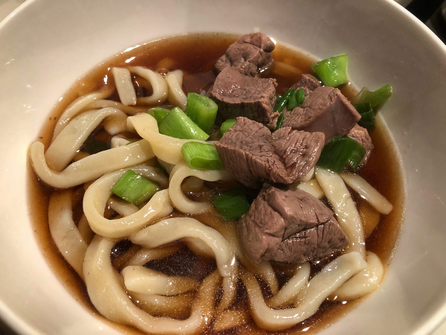 A bowl of slightly irregularly-shaped noodles in broth, topped with chunks of poached duck breast and chopped green onion