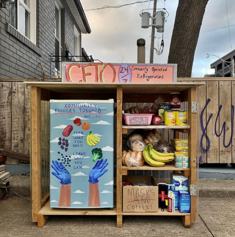 A small fridge with artwork of two hands holding up fruits and vegetables with a sign that says, "Take what you need. Leave what you can." Shelves on the side with bread, bananas, masks, and canned goods.