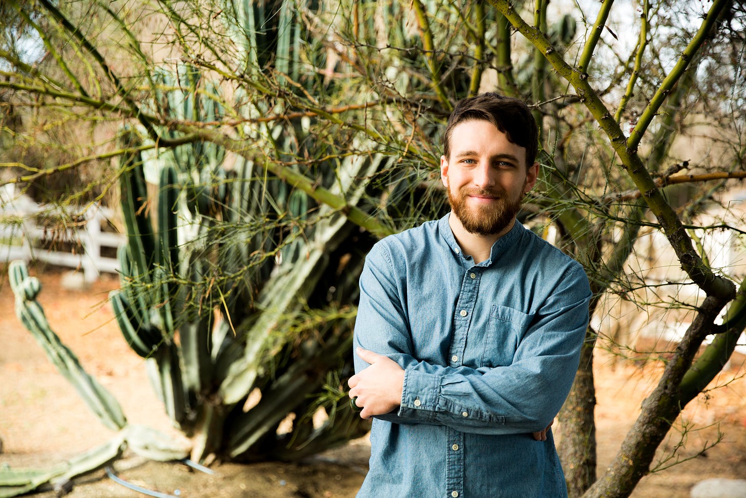a man stands in front of cactuses and greenery with a smile and arms crossed - a nice picture