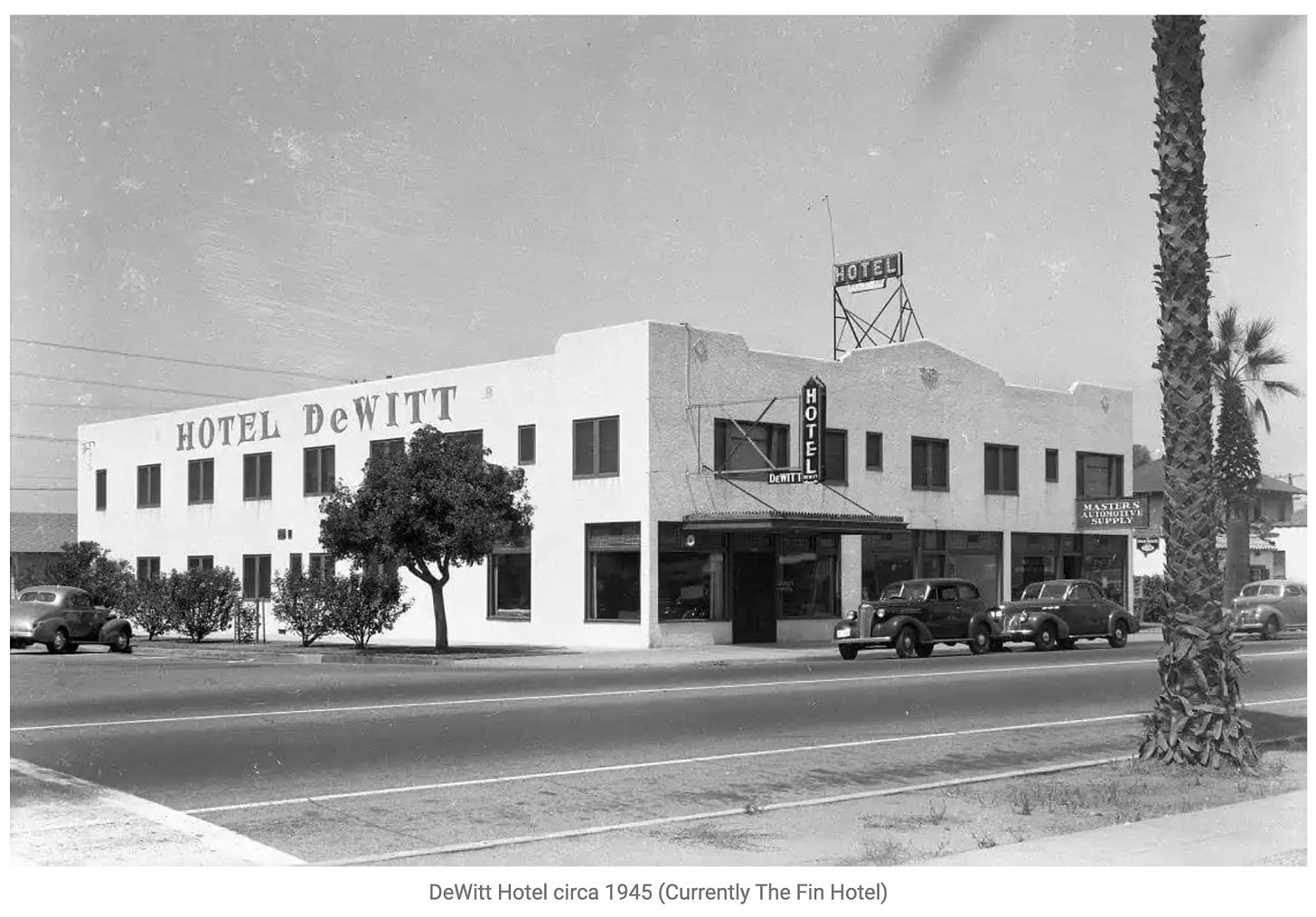 A corner view of a two-story block building with the name Hotel DeWitt painted on the side. The photo is black and white from 1945. Cars of the era are parked in front.
