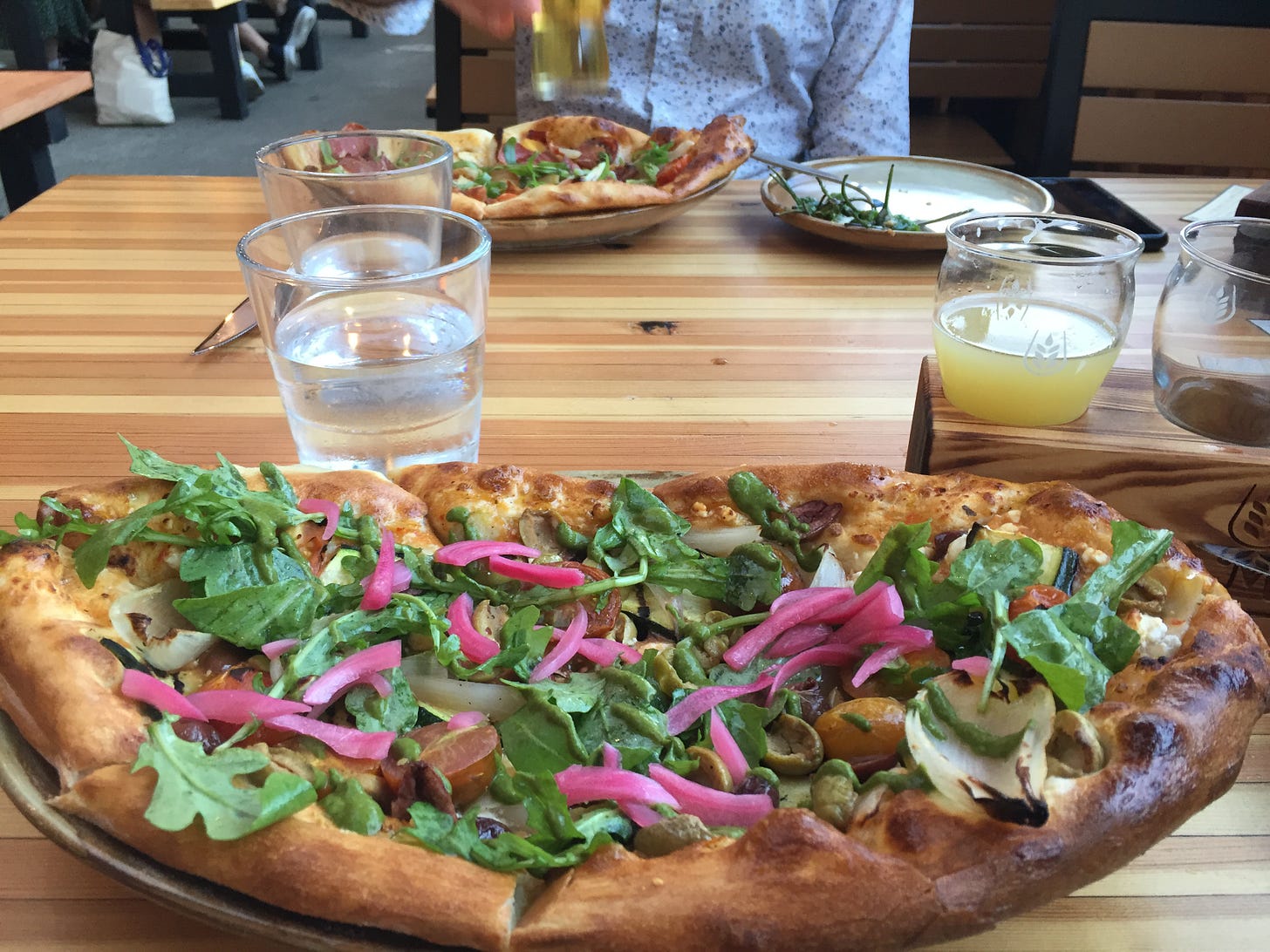 In the foreground, a flatbread pizza with pickled red onions, arugula, and olives is visible. A flight of beer in a wooden block sits behind it and to the right. In the background, another flatbread pizza is in front of Jeff, who is picking up a glass of beer.