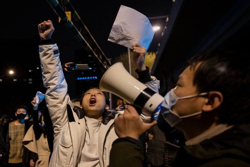 A woman with her arms raised holds up a blank white sheet of paper while a man nearby holds a megaphone to his mouth.
