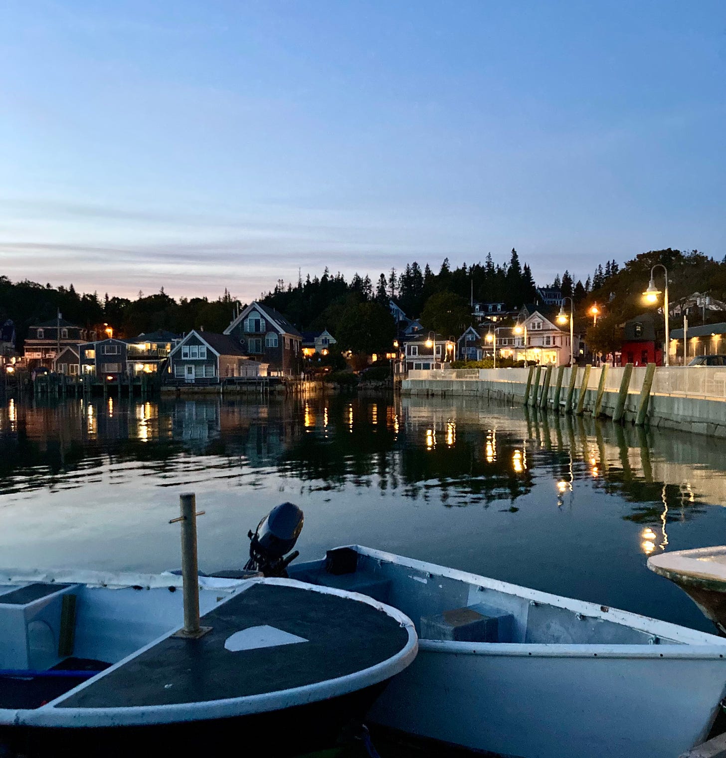 Downtown Stonington, just after sunset. There are two small boats in the water in the foreground, and yellow lights and streetlamps in the background.