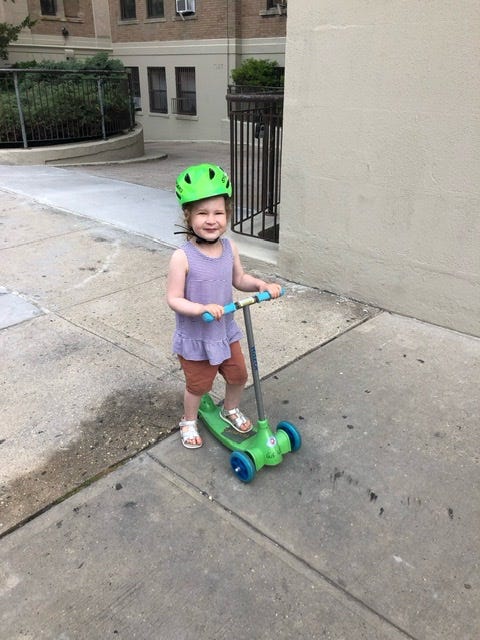 A 3-year-old girl in a green helmet and purple tank top on a green 3-wheeled scooter on a sidewalk.
