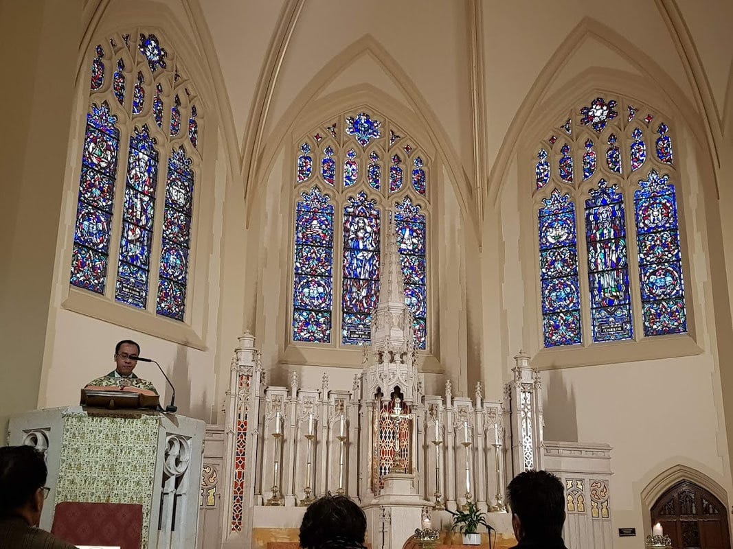 Kyrkinteriör med tre stora fönster med glasmålningar ovanför ett altarskåp i gammal stil. Till vänster en präst i en predikstol.