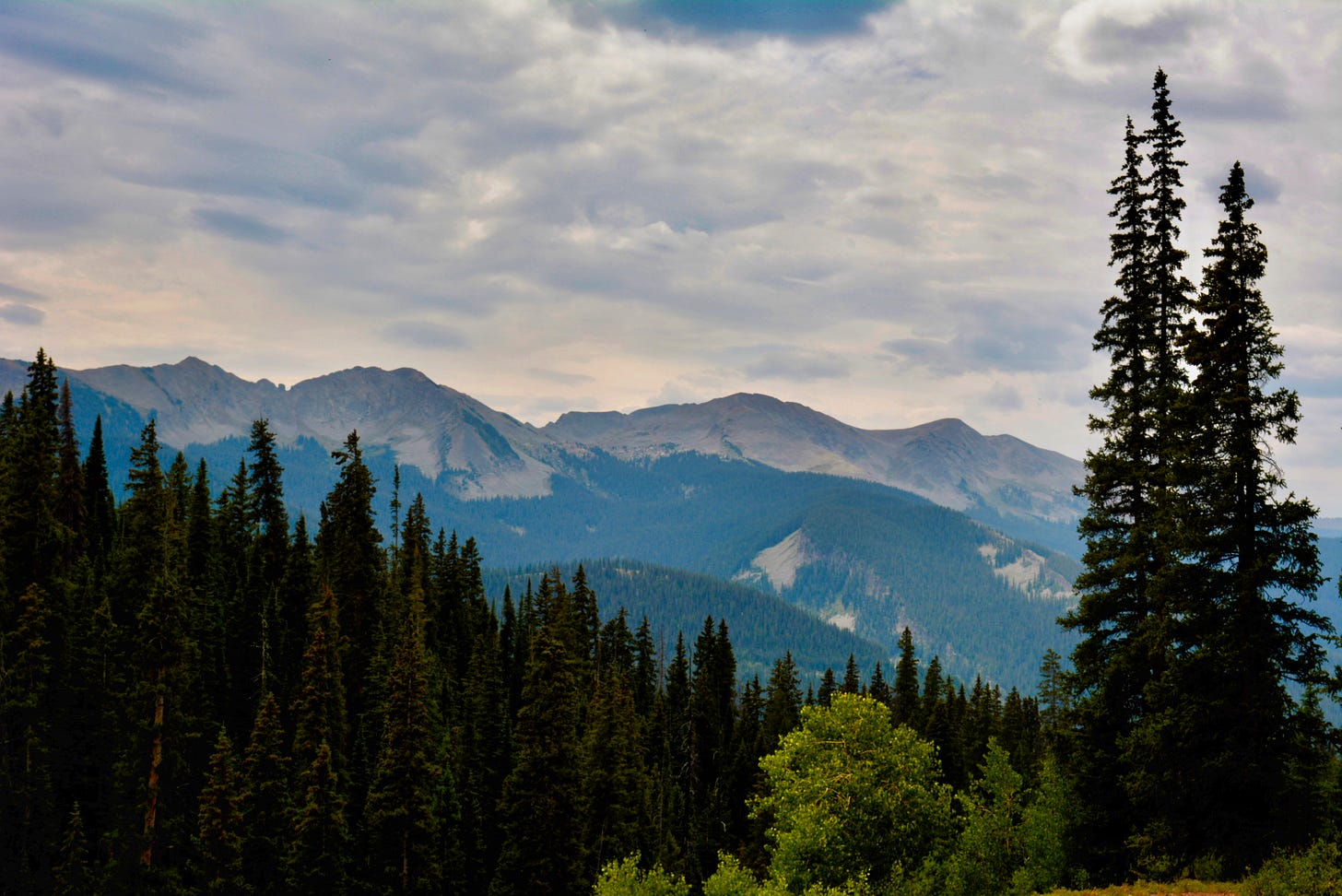 a view of the colorado mountains with trees in the foreground against a cloudy sky with specks of blue showing between the clouds