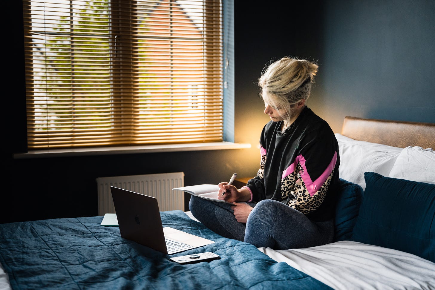 mulher loira escrevendo em um caderno sentada em cima da cama, o laptop aberto em frente a ela