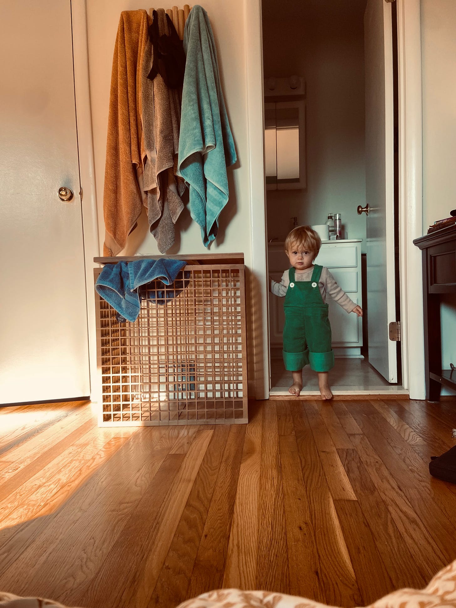 A child in green overalls is stepping over a doorway ledge into a room with white walls and a wooden floor. There is a closed door and wooden hamper to the left. There are three different colored towels hanging above the hamper while one is laying on the edge of the opening of the hamper.