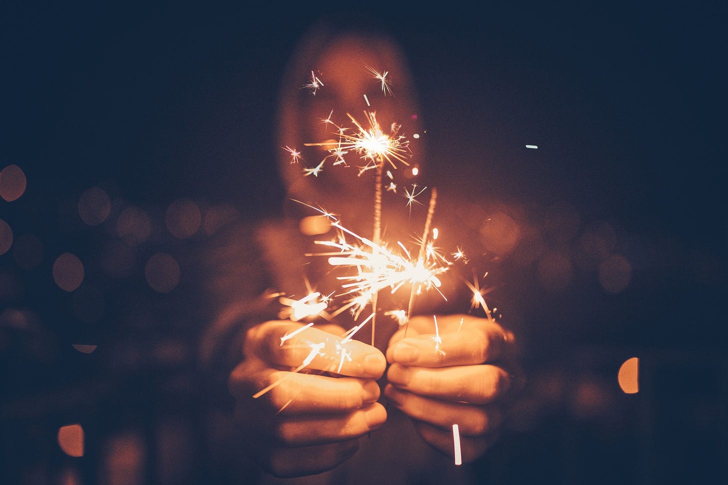 A man holds two sparklers up to the camera against a blurred background.