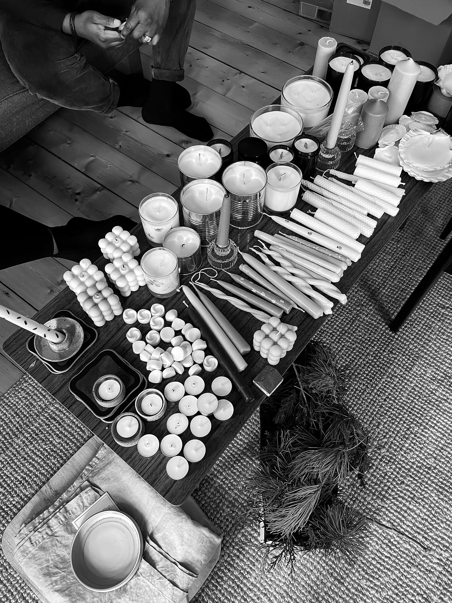 A coffee table with a wide array of different types of candles: tins, tea lights, dinner candles all laid out. There are two people's feet under the table.