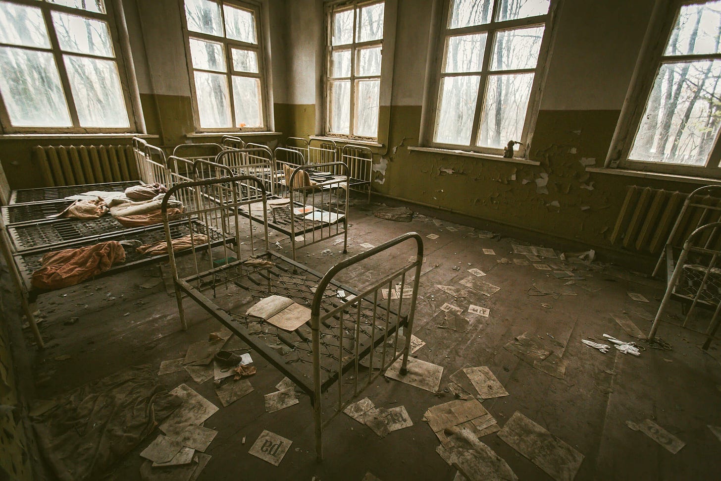 Bed frames in an abandoned room in an asylum, papers on the floor, big windows.