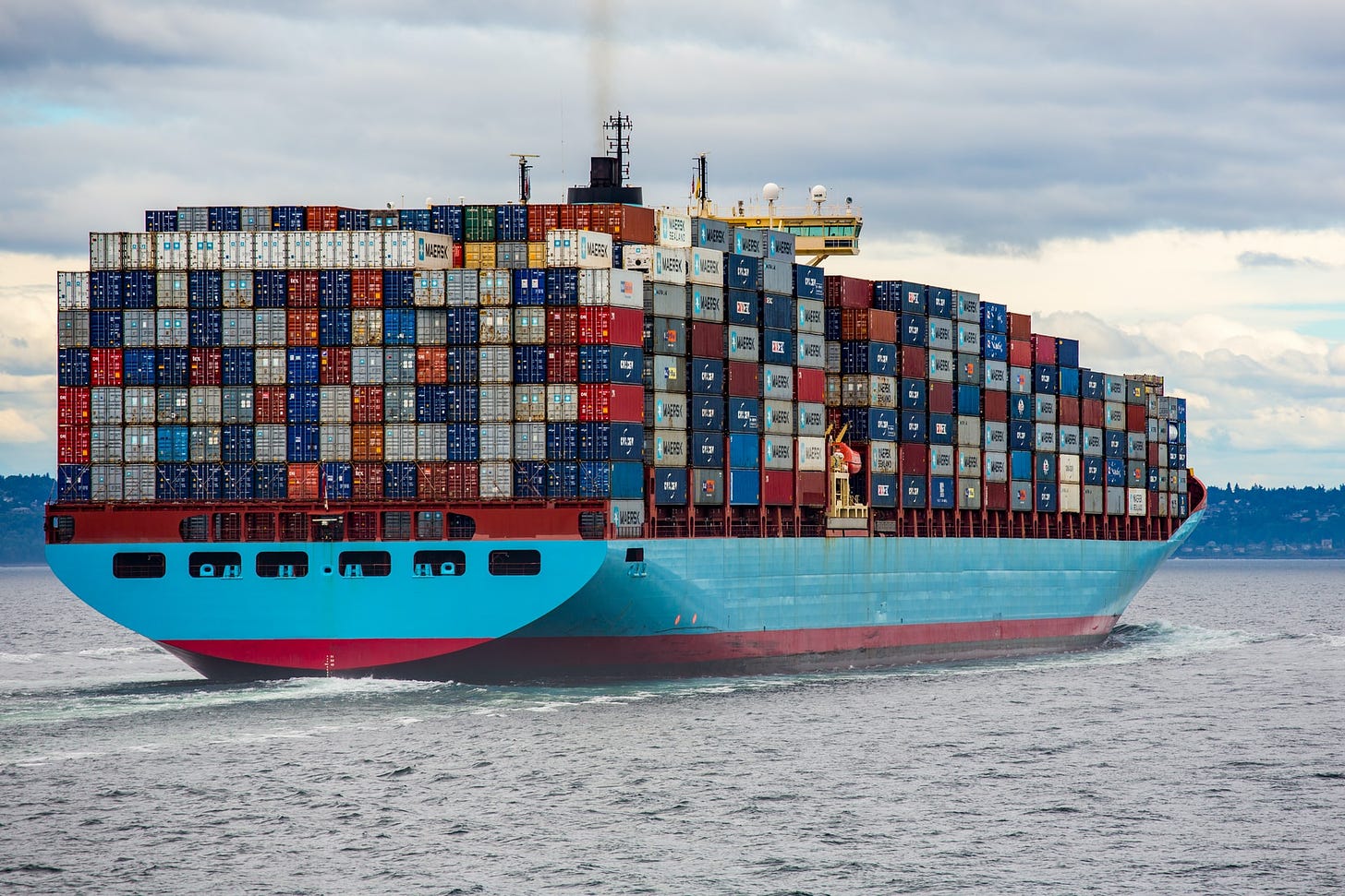 A large container ship is steaming away from the camera. The ship's base is painted sky blue, and blue, grey, red and yellow container rectangles are stacked up to eight containers high. A slight bit of smoke comes from a pipe floating up into a cloudy sky.