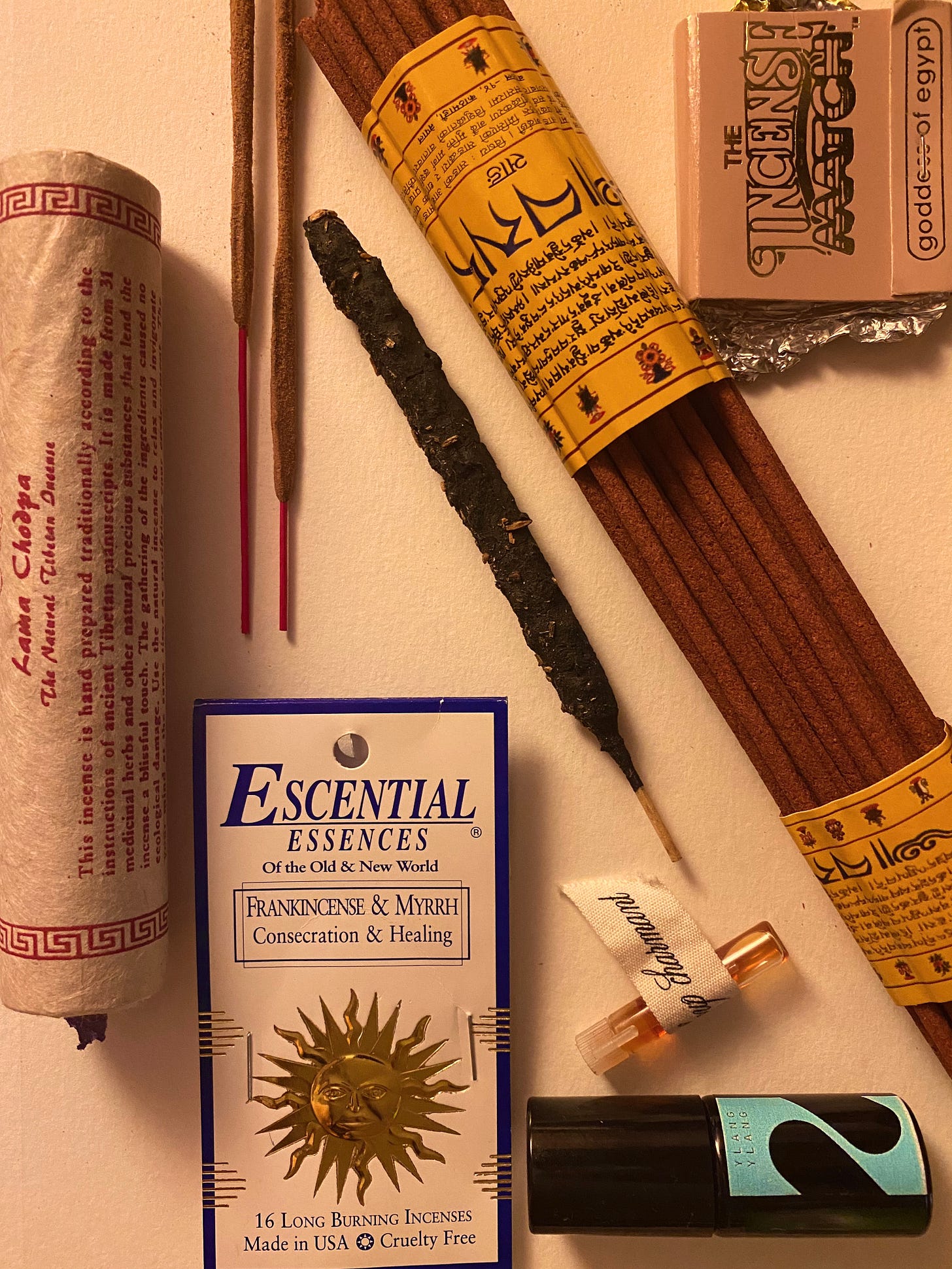 An array of different incense and two small perfume bottles of ylang ylang and a musky sweet scent named "loup charmant." One of the incense is a brand "Escential Essences," another is an incense matchbook named "goddess of egypt," and another is a traditional tibetan hand rolled incense. the image is warm in tone 