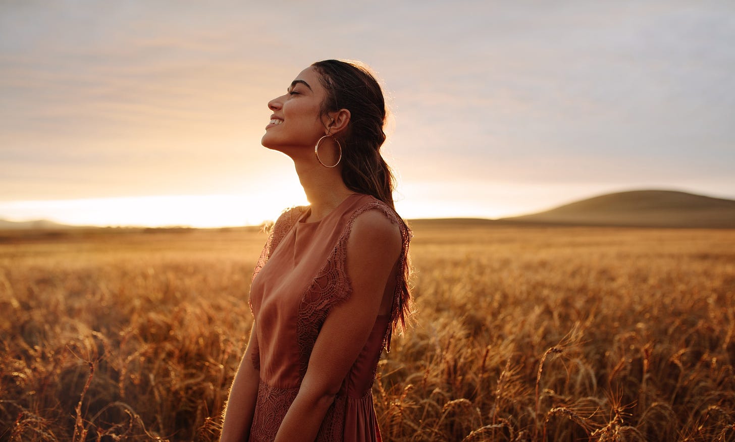 A woman with long dark hair smiles in a field as the sun sets.