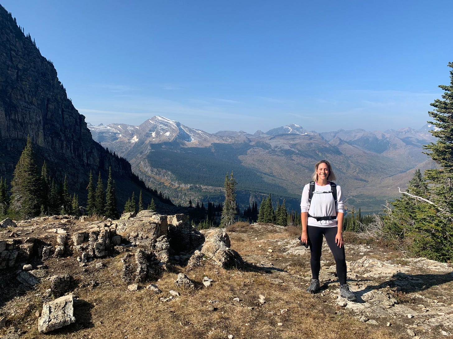 A woman in hiking boots, black workout pants, a white longsleeve shirt, and a backup smiles at the camera on top of a mountain peak.