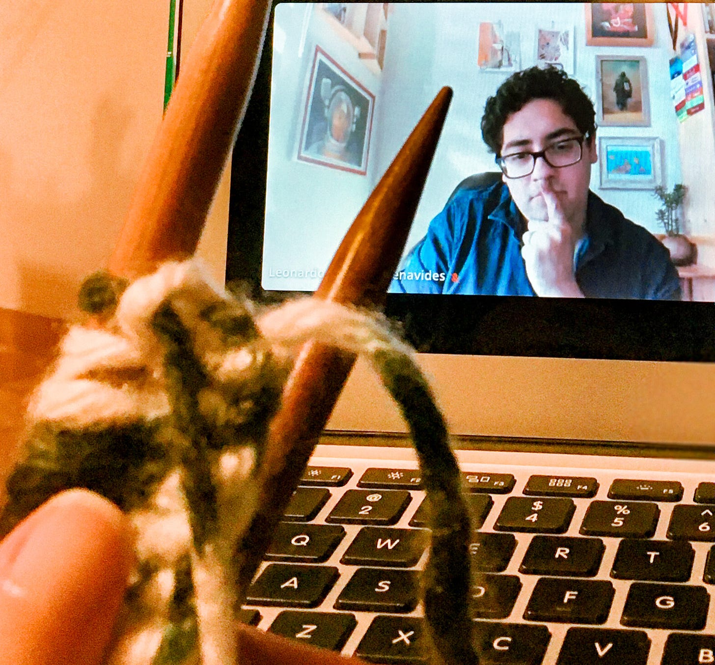 Na tela de computador, se observa um homem moreno, de óculos, com semblante pensativo. Em primeiro plano, agulhas de tricô e um teclado.