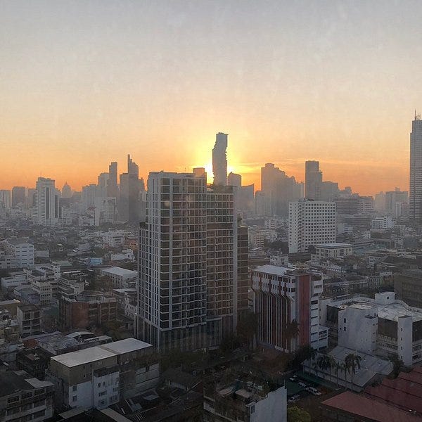 A city skyline view at sunrise. Busy buildings at the front and a wobbly tall building just infant of the rising sun, and a sky of oranges fading up to light blue. 